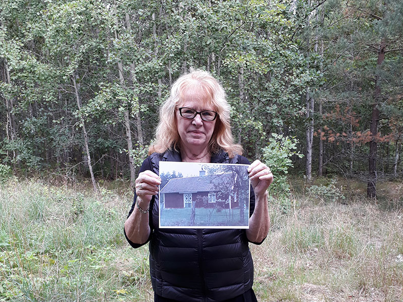 Traveler hold a photo of the cottage her ancestor lived in before he emigrated. She stand on the land where the cottage where.